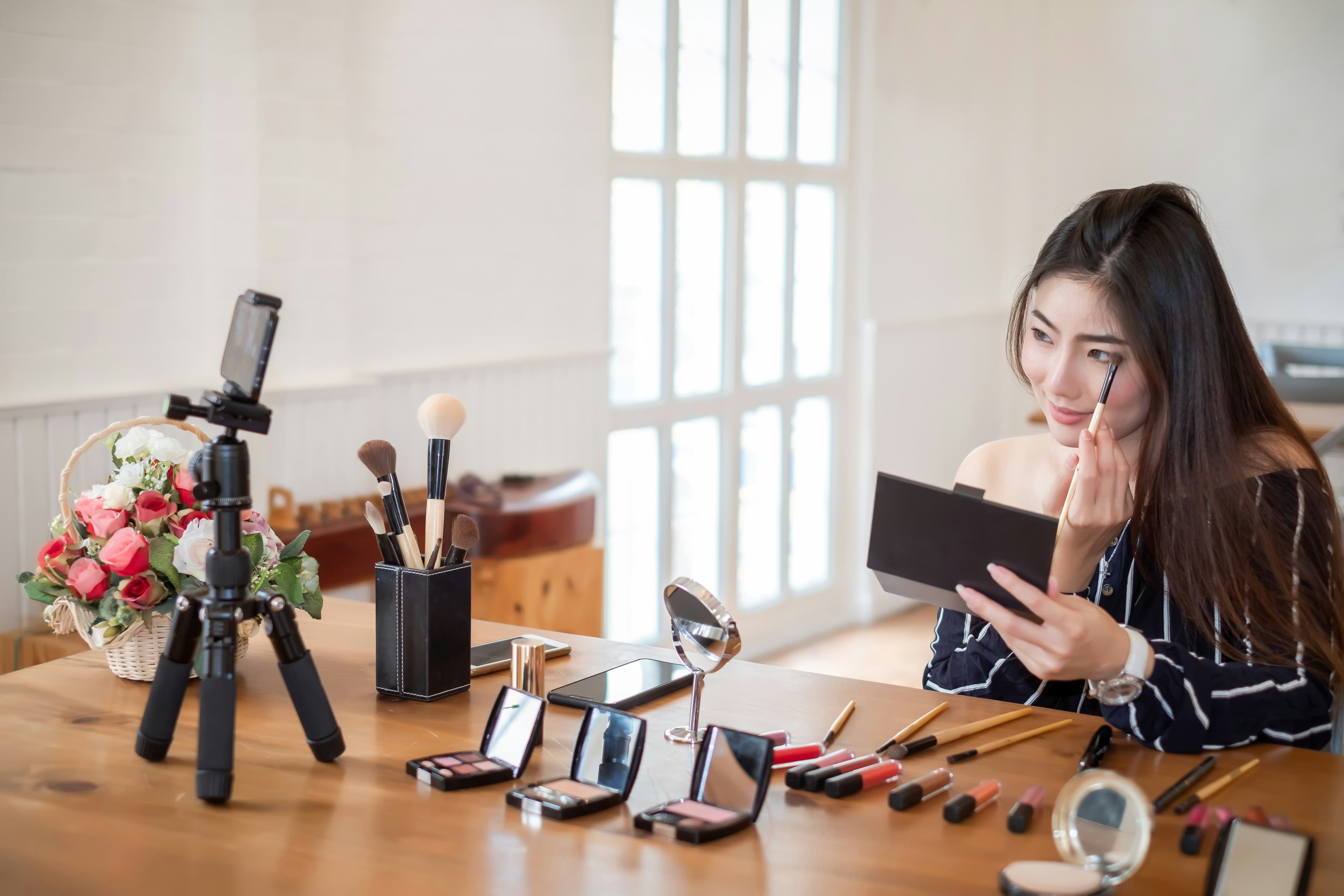 A beauty creator filming a makeup review at a tripod-mounted phone
